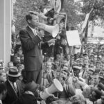 Attorney General Robert F. Kennedy speaking to a crowd of African Americans and whites through a megaphone outside the Justice Department; sign for Congress of Racial Equality is prominently displayed.